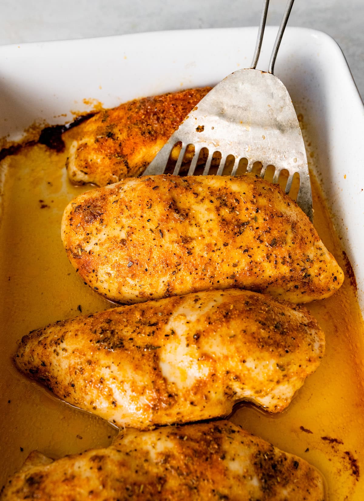 Four baked chicken breasts in a baking dish with a metal spatula lifting one up.