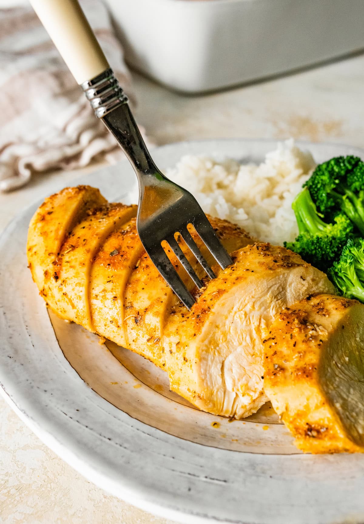 A baked chicken breast cut into slices on a plate with a side of broccoli and rice. There's a fork sticking up in one piece of chicken.