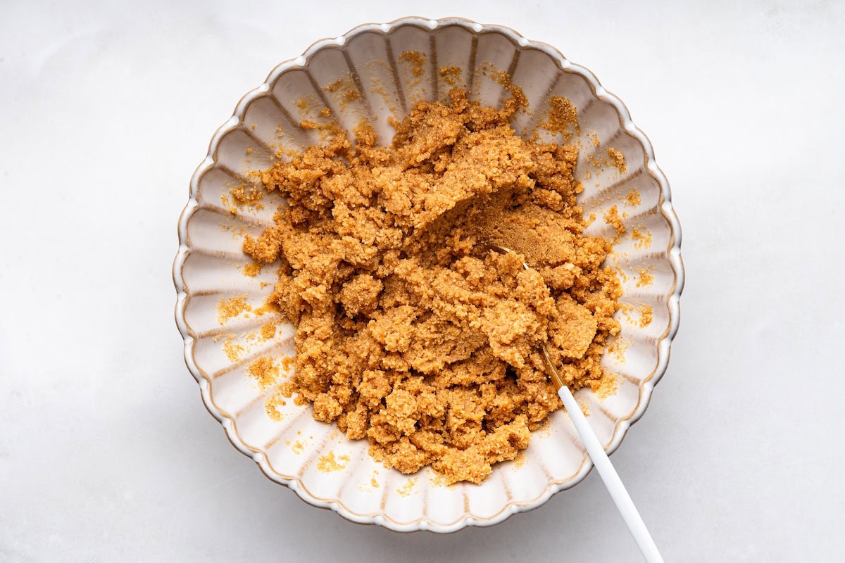 Graham cracker crumbs and maple syrup mixed together in a small mixing bowl with a white spoon.