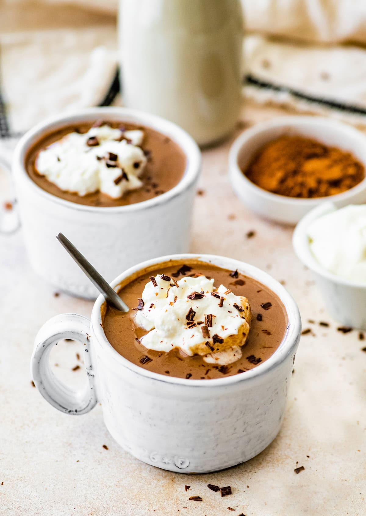 Two protein hot chocolates white ceramic mugs topped with whip cream and shaved chocolate. A silver spoon is in both of the mugs.