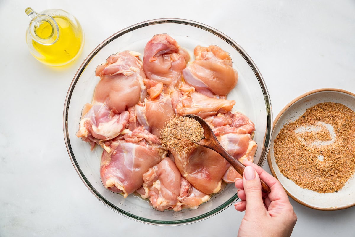 A womans hand uses a wooden spoon to add brown sugar to a large glass bowl of raw boneless chicken thighs.