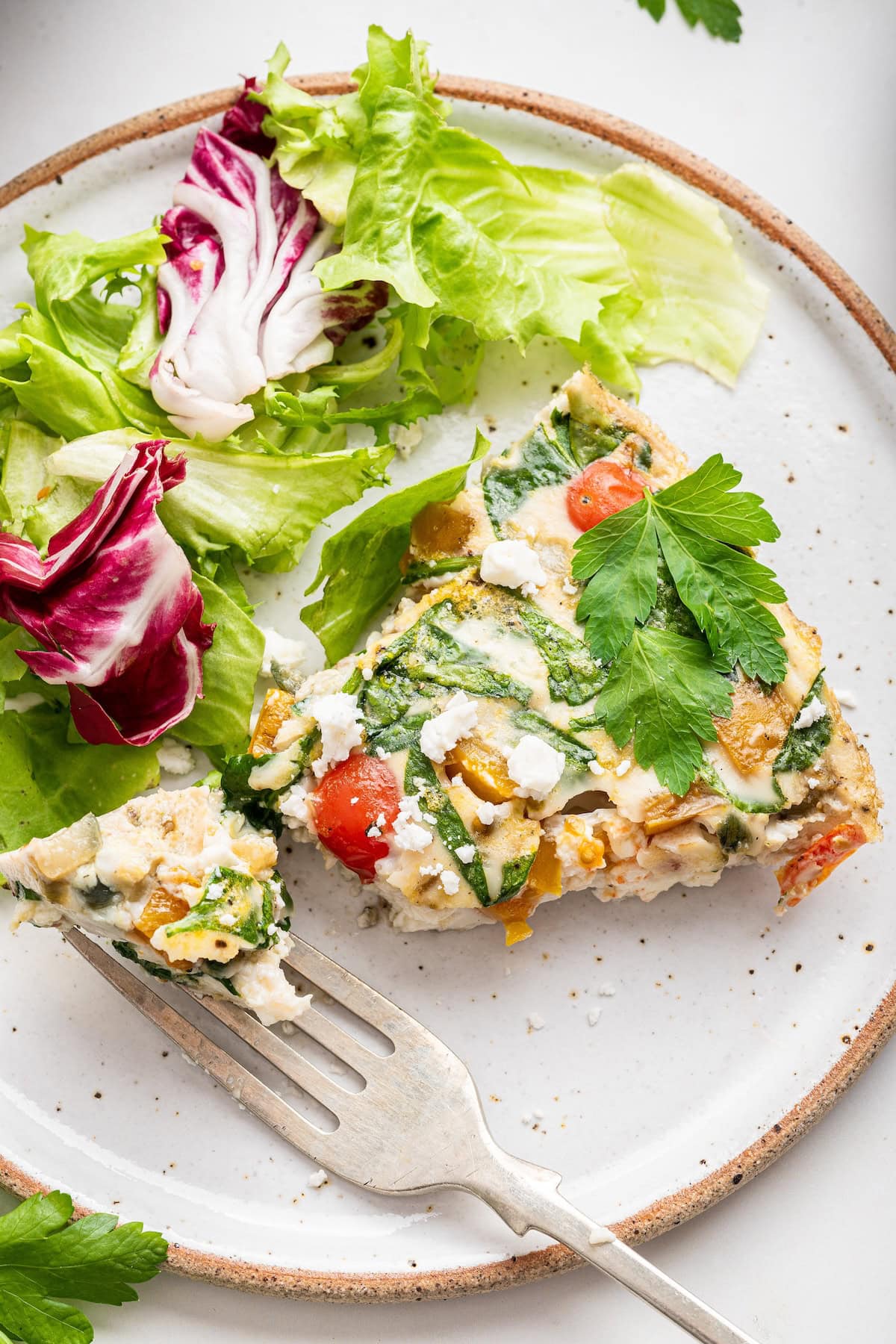 A slice of egg white frittata on a white plate with a side salad. There is a fork taking a portion of the egg white frittata on the plate.
