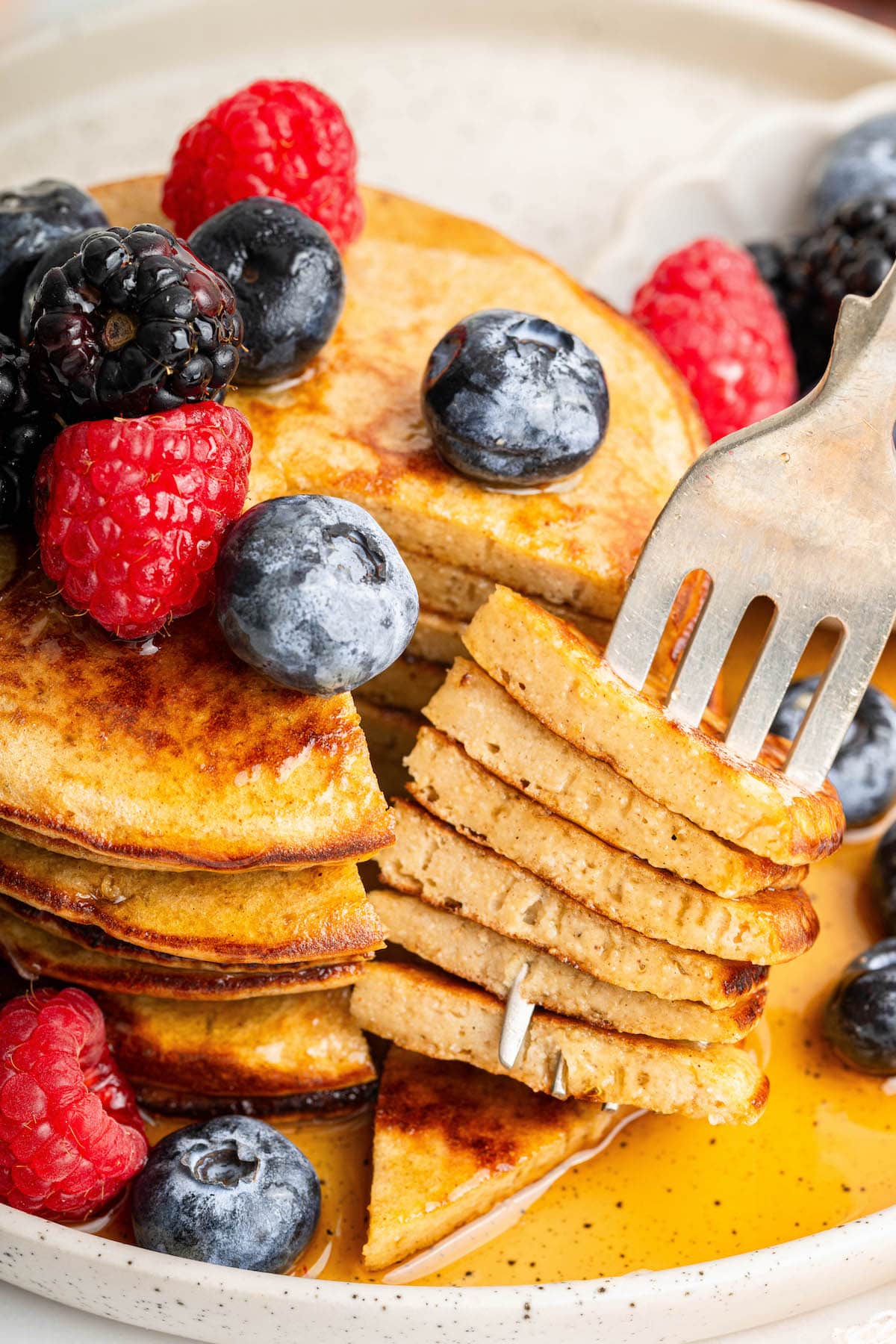 Six greek yogurt pancakes stacked on a plate topped with maple syrup, fresh blueberries, blackberries and raspberries. A fork is removing a bite from the stack.