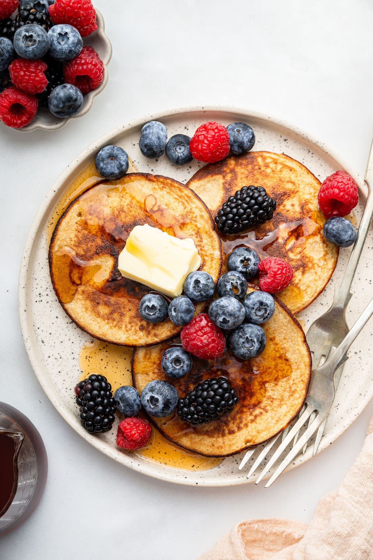 Three greek yogurt pancakes on a plate topped with butter, maple syrup and fresh blueberries, raspberries and blackberries.