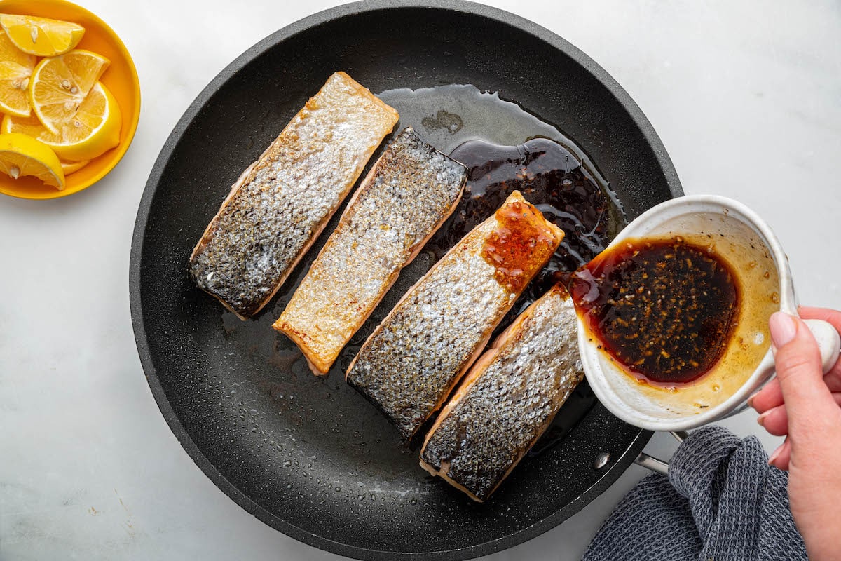 A woman's hand pours a honey based sauce over four salmon filets that are cooking in a skillet.