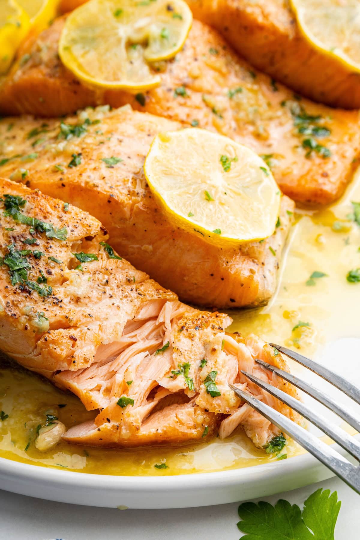 A fork digging into a salmon filet on a plate alongside three other salmon filets that are garnished with lemon wedges and fresh herbs.