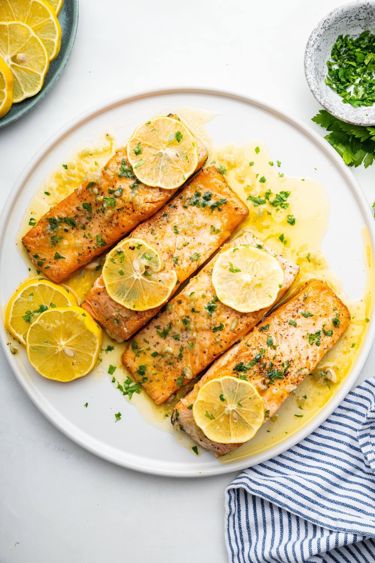 Four salmon filets covered in garlic butter on a white plate, garnished with fresh herbs and lemon wedges.