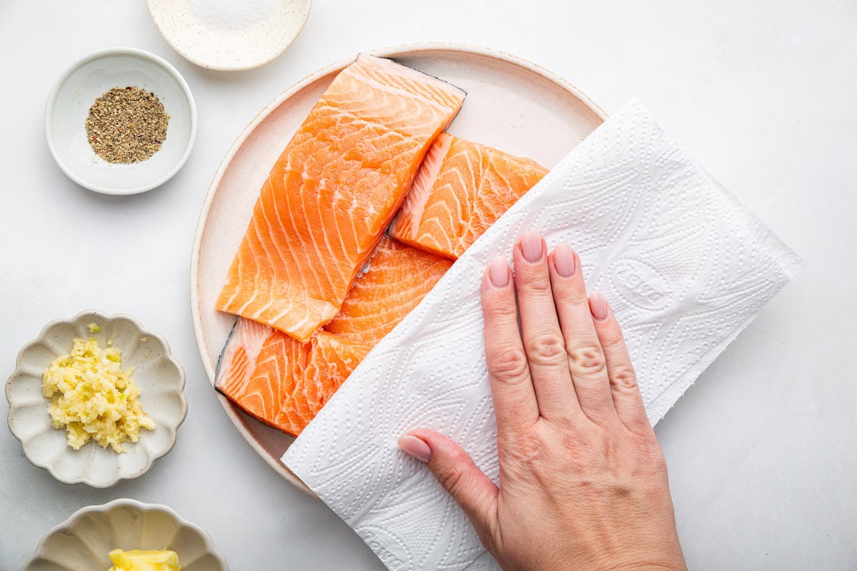 A womans hand uses a paper towel to pat dry raw salmon filets on a white plate. There are small dishes of garlic, butter, salt, and pepper to the side of the plate.