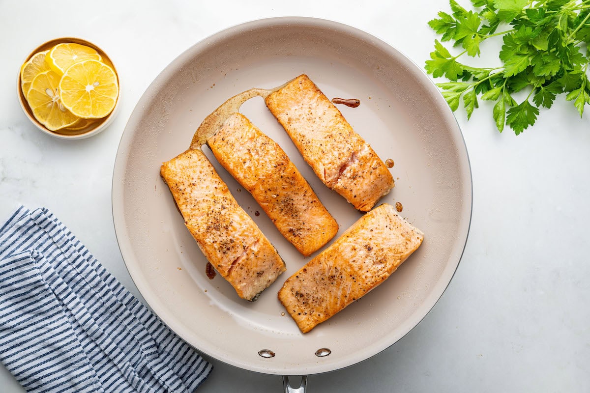 Four pan seared salmon filets in a tan skillet.