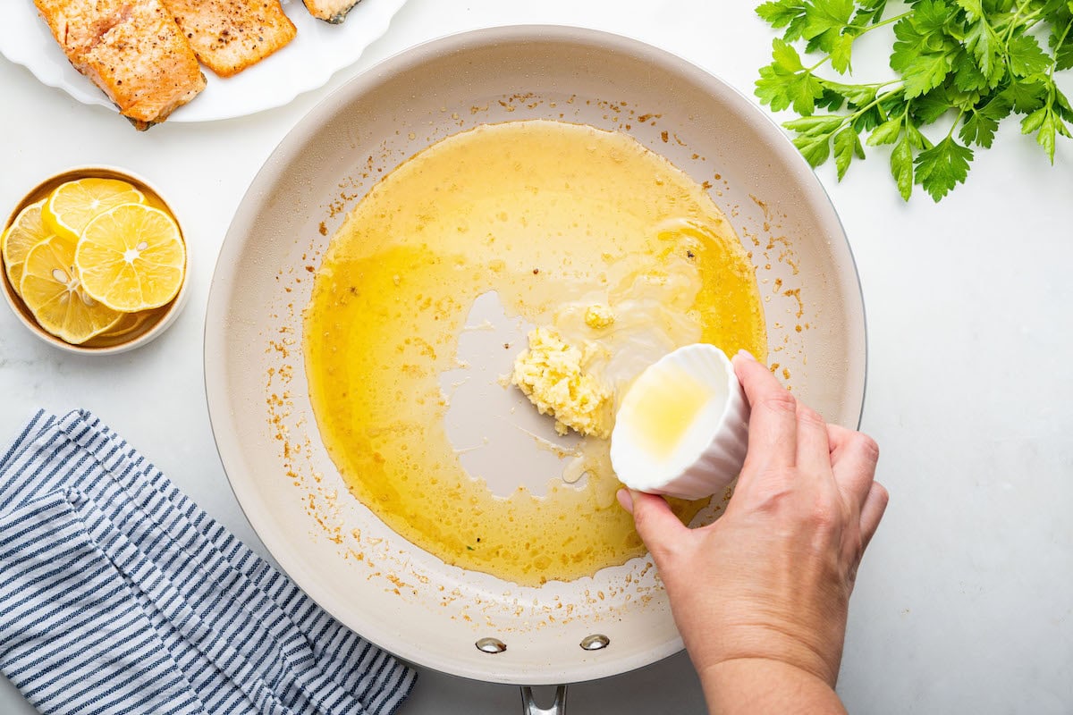 A womans hand pours melted butter in a pan along with minced garlic.