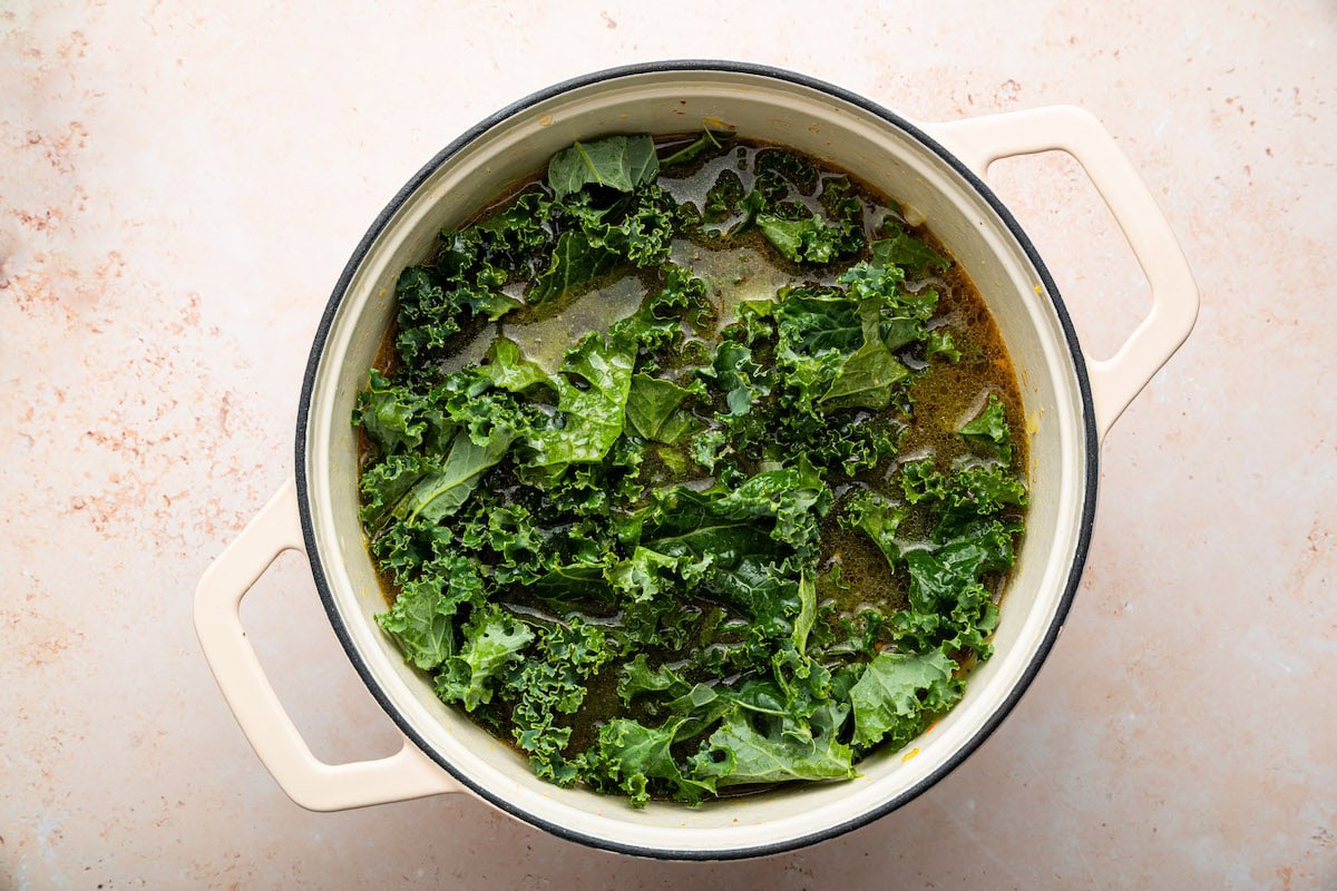 Chopped kale being added to the sausage and kale soup in a pot.