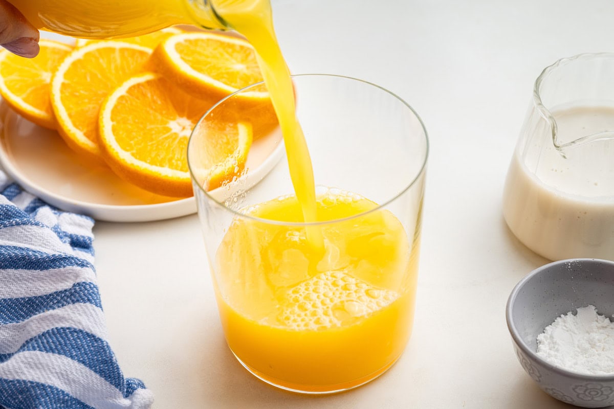 Orange juice being poured into a glass. Orange slices on a plate are in the background.