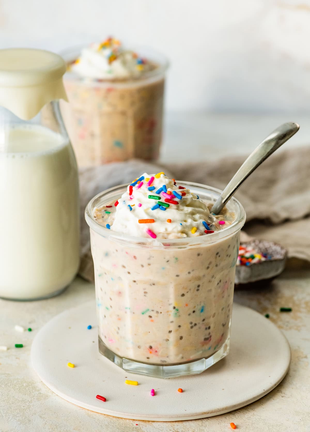 A glass jar filled with birthday cake overnight oats, topped with whipped cream and colorful sprinkles, with a bottle of milk and another jar of oats in the background.