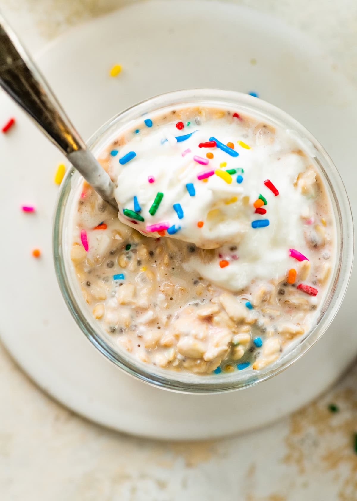 Birthday cake overnight oats in a glass jar with a spoon, showing creamy oats, whipped topping, and rainbow sprinkles.
