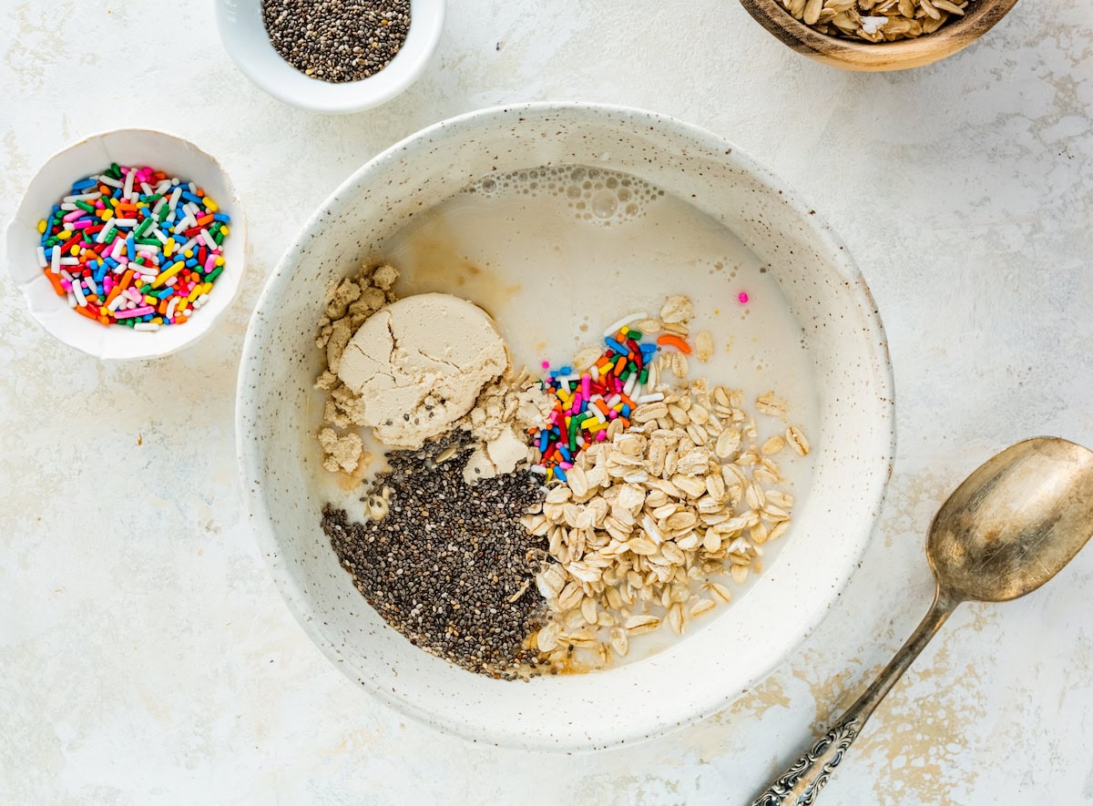 A bowl filled with oats, chia seeds, protein powder, almond milk, and colorful sprinkles, ready to be mixed for birthday cake overnight oats.