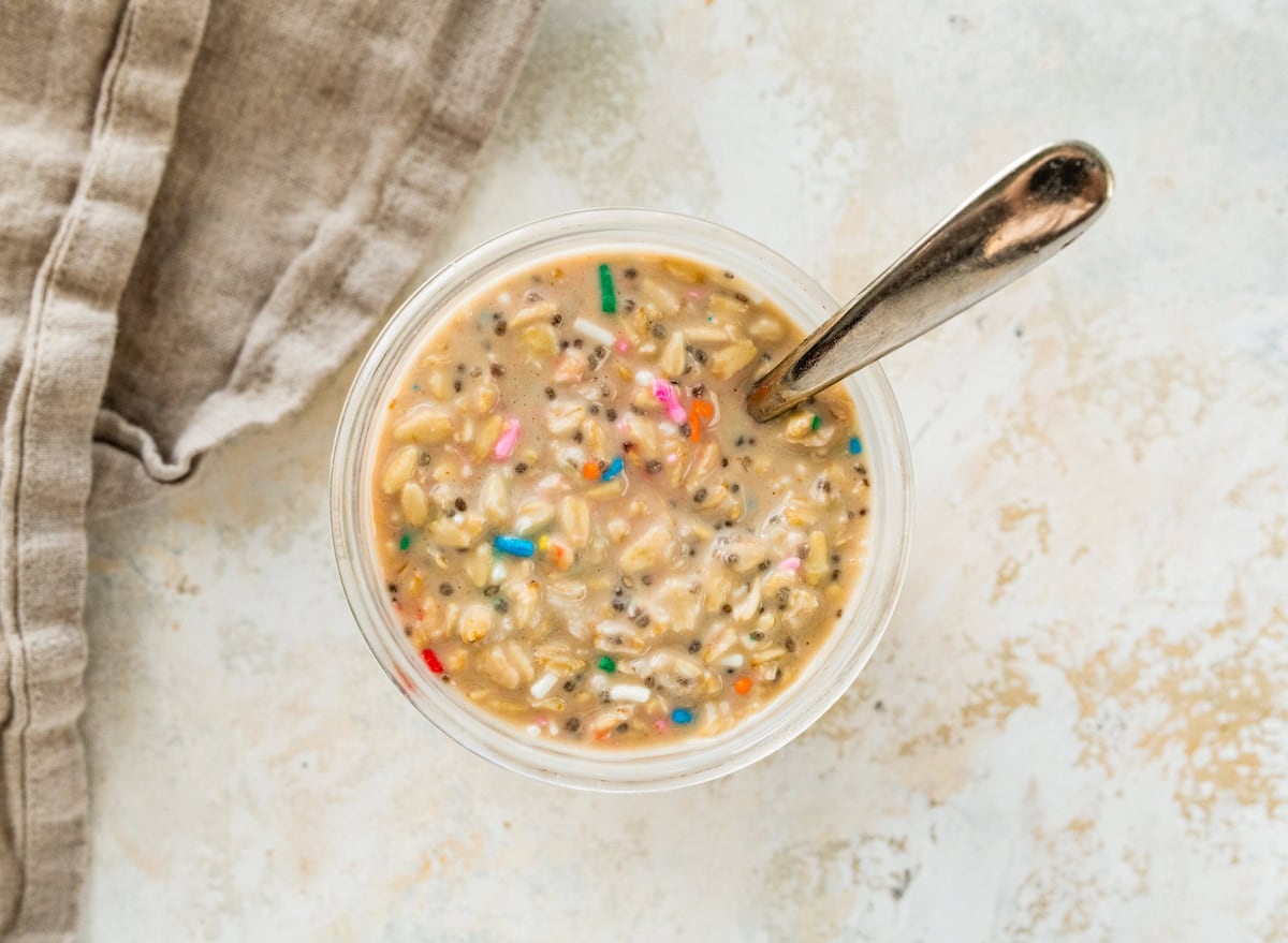 Glass jar of birthday cake overnight oats with a spoon, showing softened oats, chia seeds, and rainbow sprinkles.