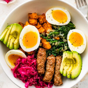 A white bowl serving two hard boiled eggs split open with avocado slices, sausage, sweet potatoes, kale, and sauerkraut.