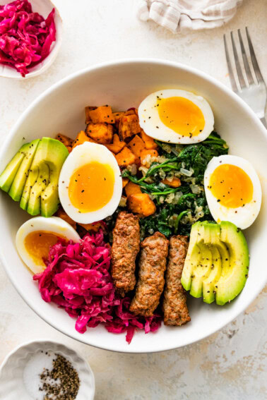A white bowl serving two hard boiled eggs split open with avocado slices, sausage, sweet potatoes, kale, and sauerkraut.