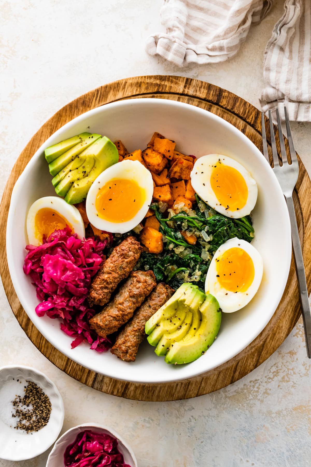 A serving of a savory breakfast bowl with hard boiled eggs, sweet potatoes, avocado slices, turkey sausage, sauerkraut, and cooked kale.