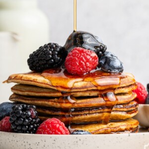 Maple syrup being poured on top of a stack of six greek yogurt pancakes on a plate. They're topped with fresh blueberries, blackberries and raspberries.