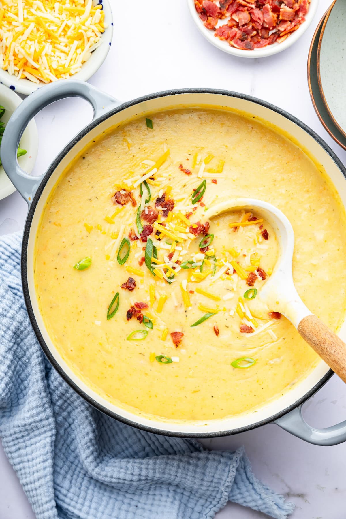 A ladle scooping out a spoonful of potato soup from a dutch oven pot. The soup is topped with bacon bits, green onion and shredded cheese.