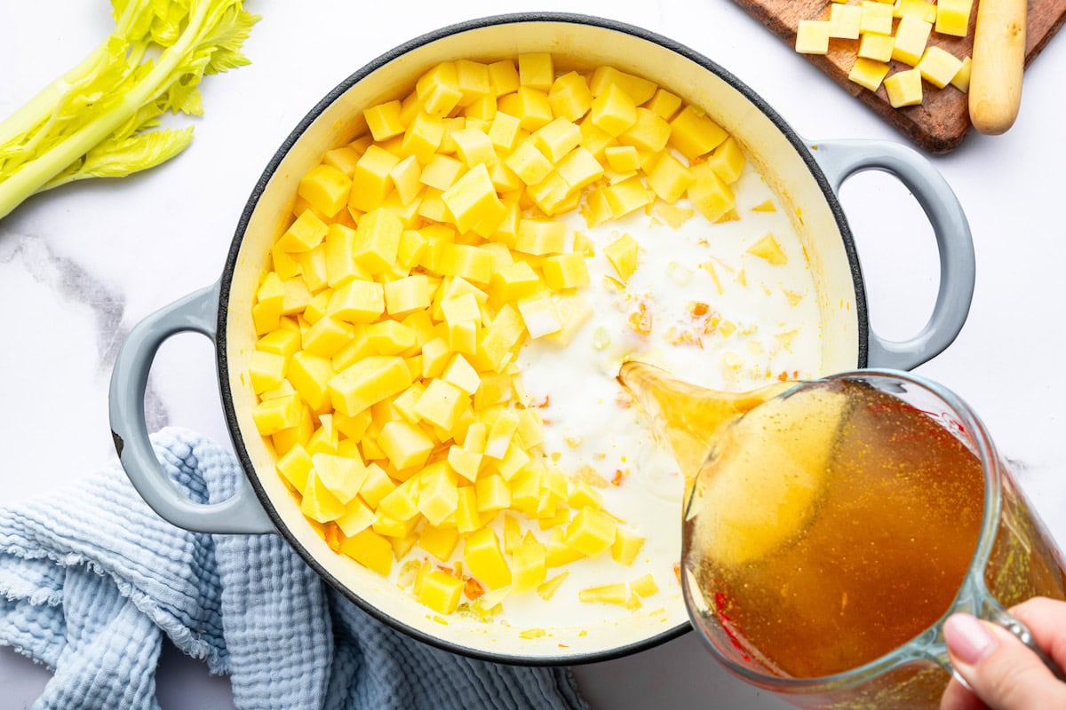A woman's hand pouring vegetable broth into a dutch oven pot filled with chopped potatoes, milk and sautéed vegetables.