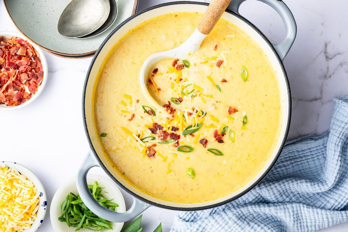 A ladle scooping out a spoonful of potato soup from a dutch oven pot. The soup is topped with bacon bits, green onion and shredded cheese.