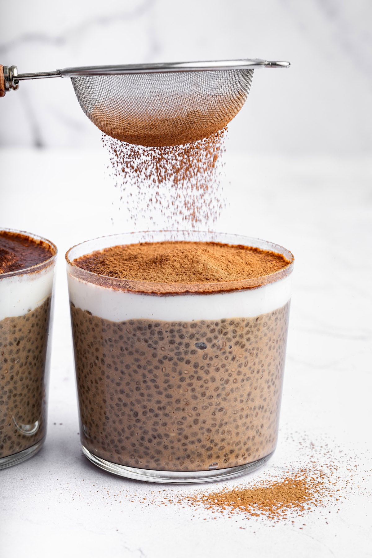 Two glass cups of tiramisu chia pudding near one another. A small metal sifter is being used to add cocoa powder on top of the pudding.