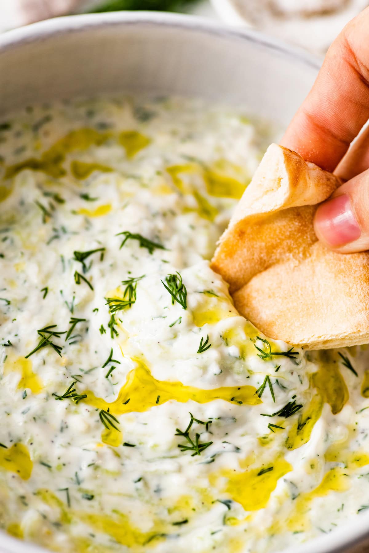 A woman's hand dipping a piece of pita bread in tzatziki sauce. The sauce is topped with fresh dill and olive oil.