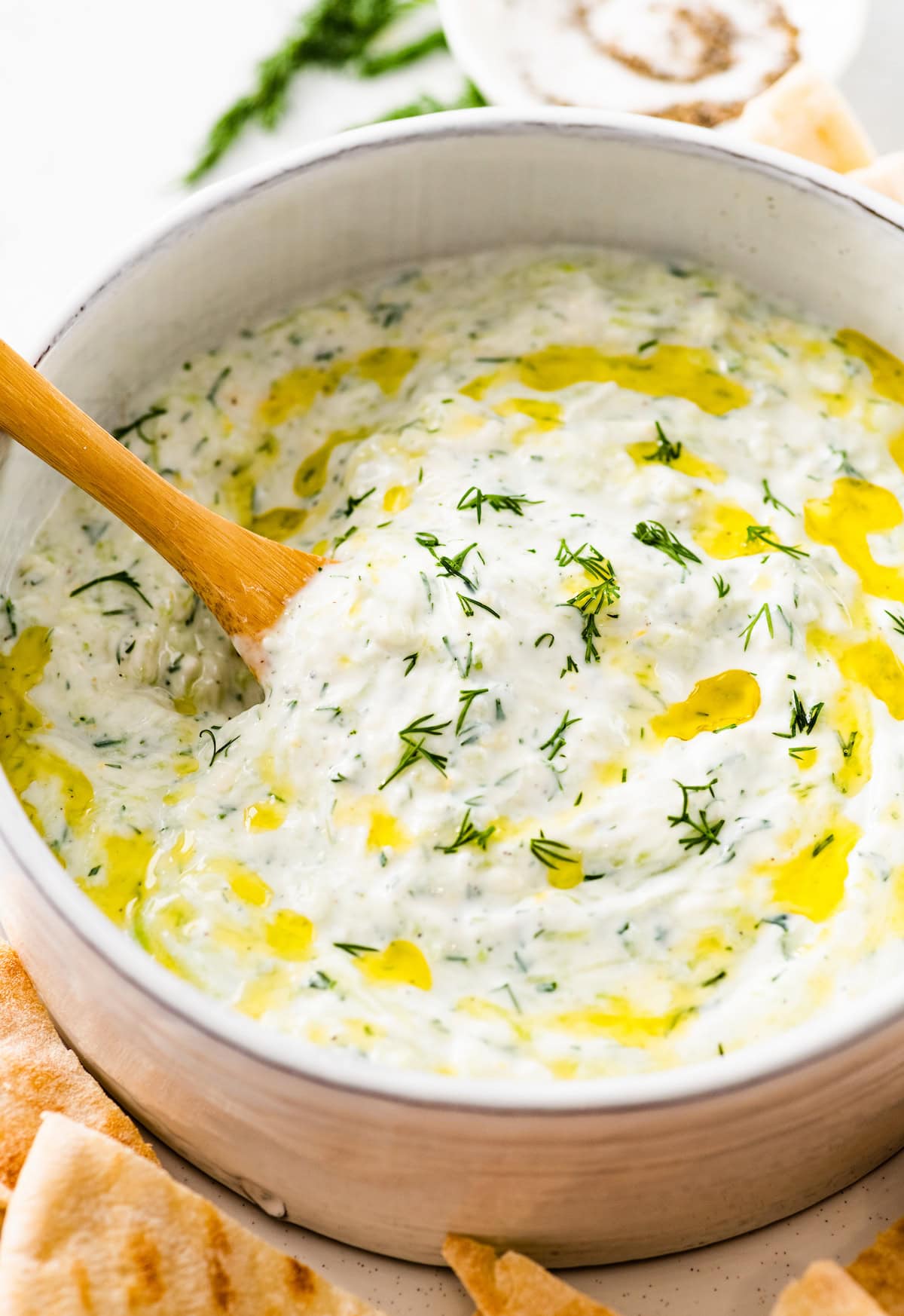 A white bowl of tzatziki topped with fresh dill and olive oil. There is a wooden spoon in the bowl and the bowl is surrounded by pita bread.