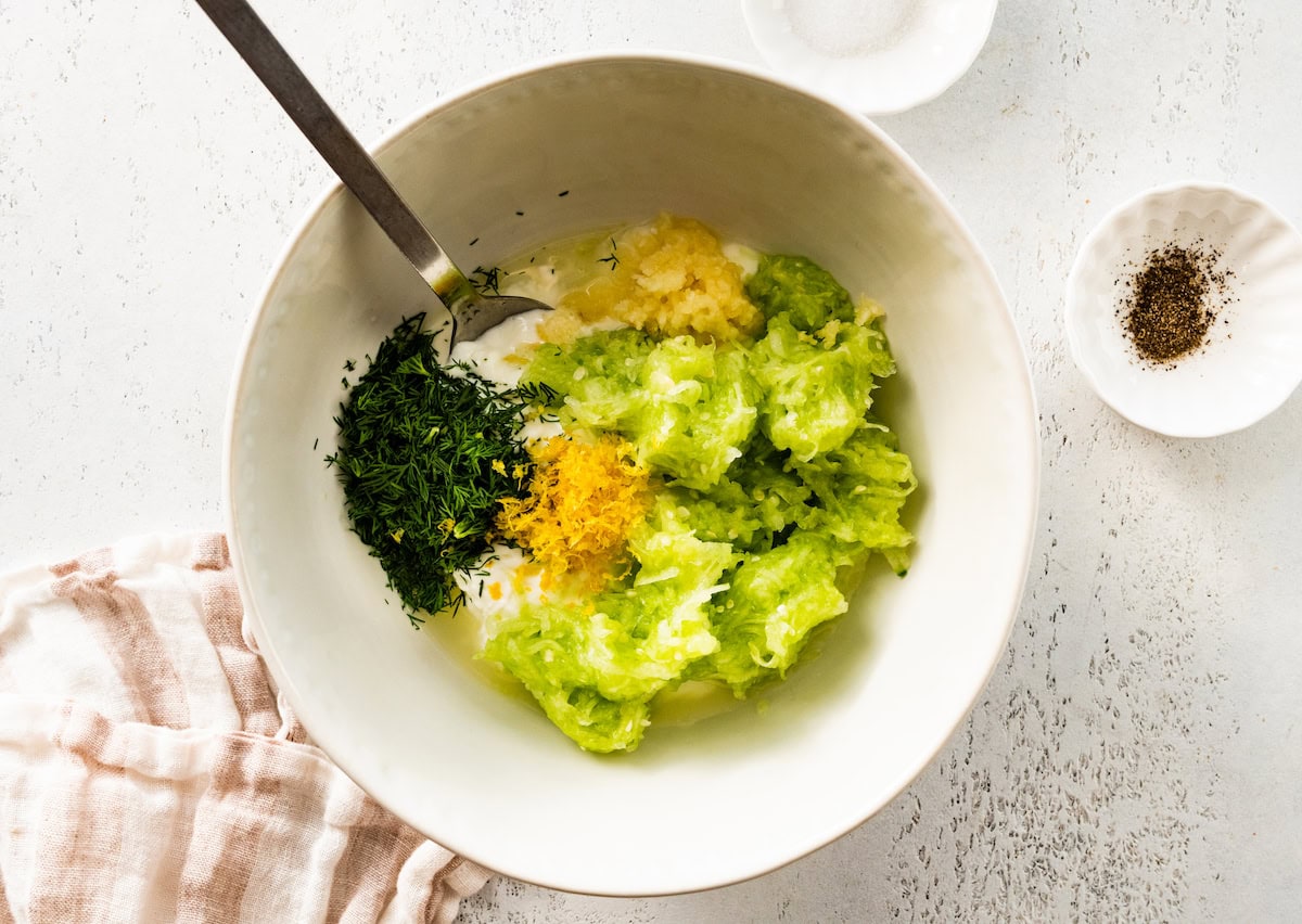 All of the tzatziki ingredients in a white mixing bowl: garlic, grated cucumber, greek yogurt, olive oil and fresh dill. There is a silver spoon in the bowl.