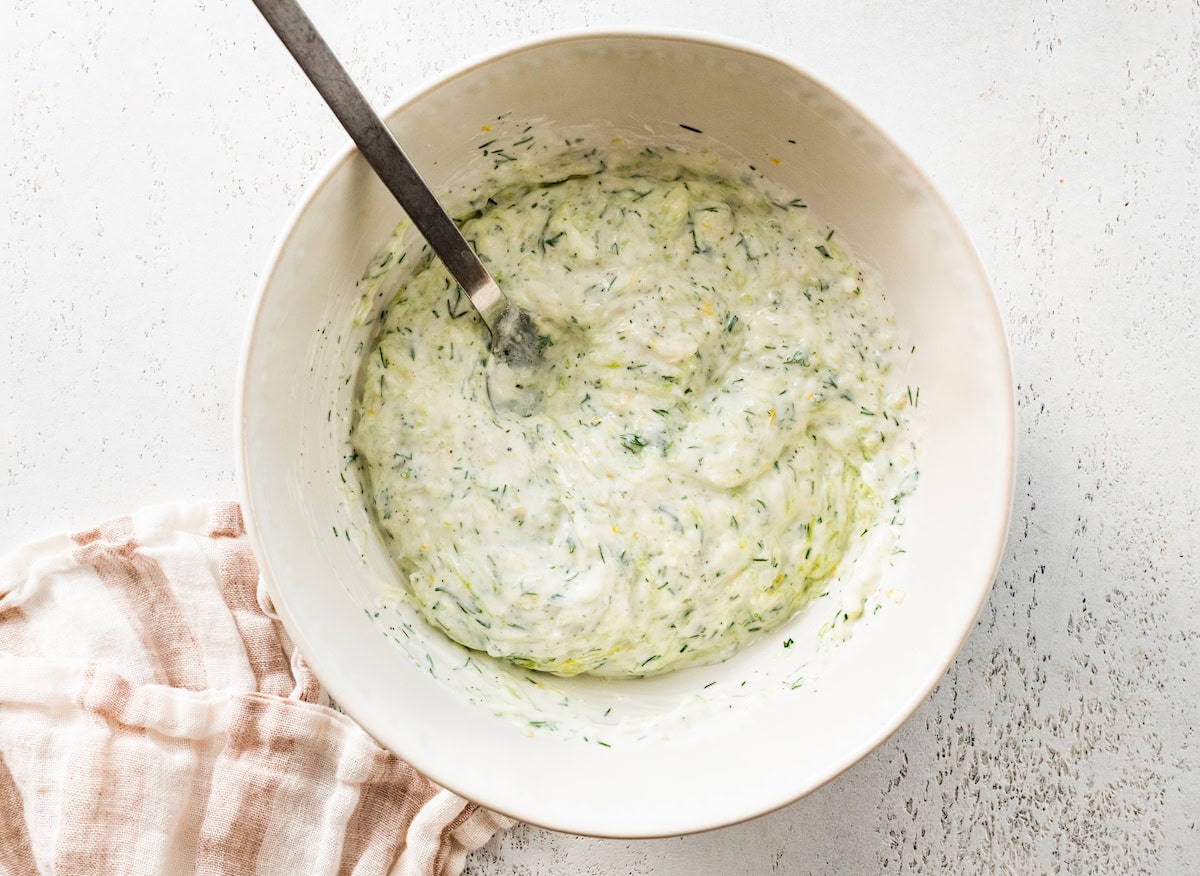 The mixed tzatziki sauce in a white mixing bowl. There is a silver spoon in the bowl.