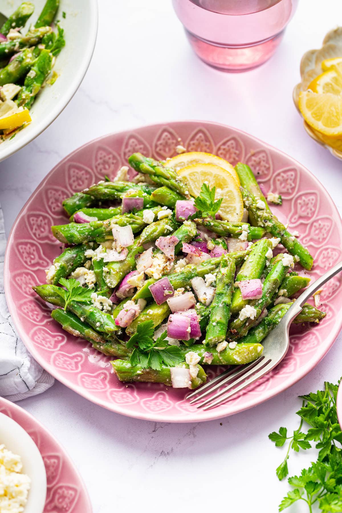 A serving of asparagus salad on a pink patterned plate with a fork, featuring chopped asparagus, red onion, feta, fresh parsley, and lemon slices.