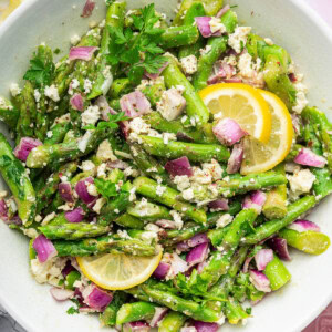 An asparagus salad served in a white bowl with feta, red onion, and lemon wedges.