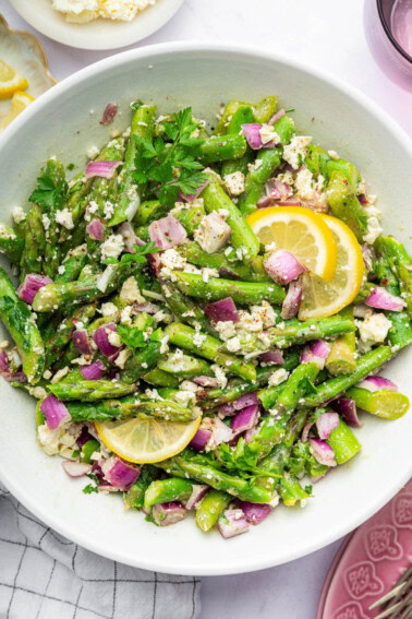 An asparagus salad served in a white bowl with feta, red onion, and lemon wedges.