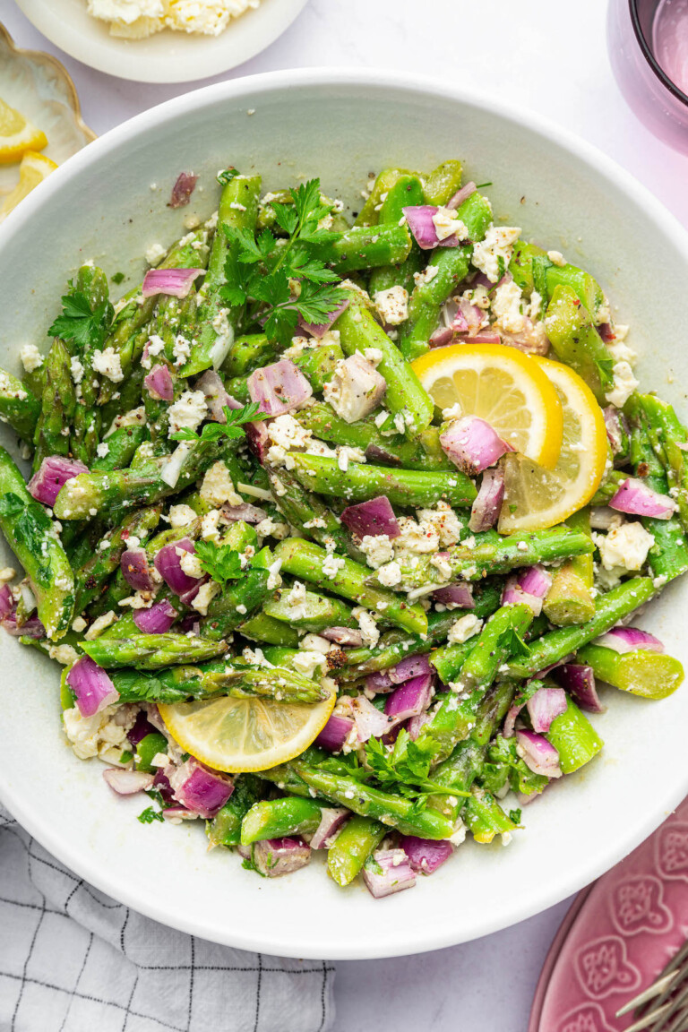 An asparagus salad served in a white bowl with feta, red onion, and lemon wedges.