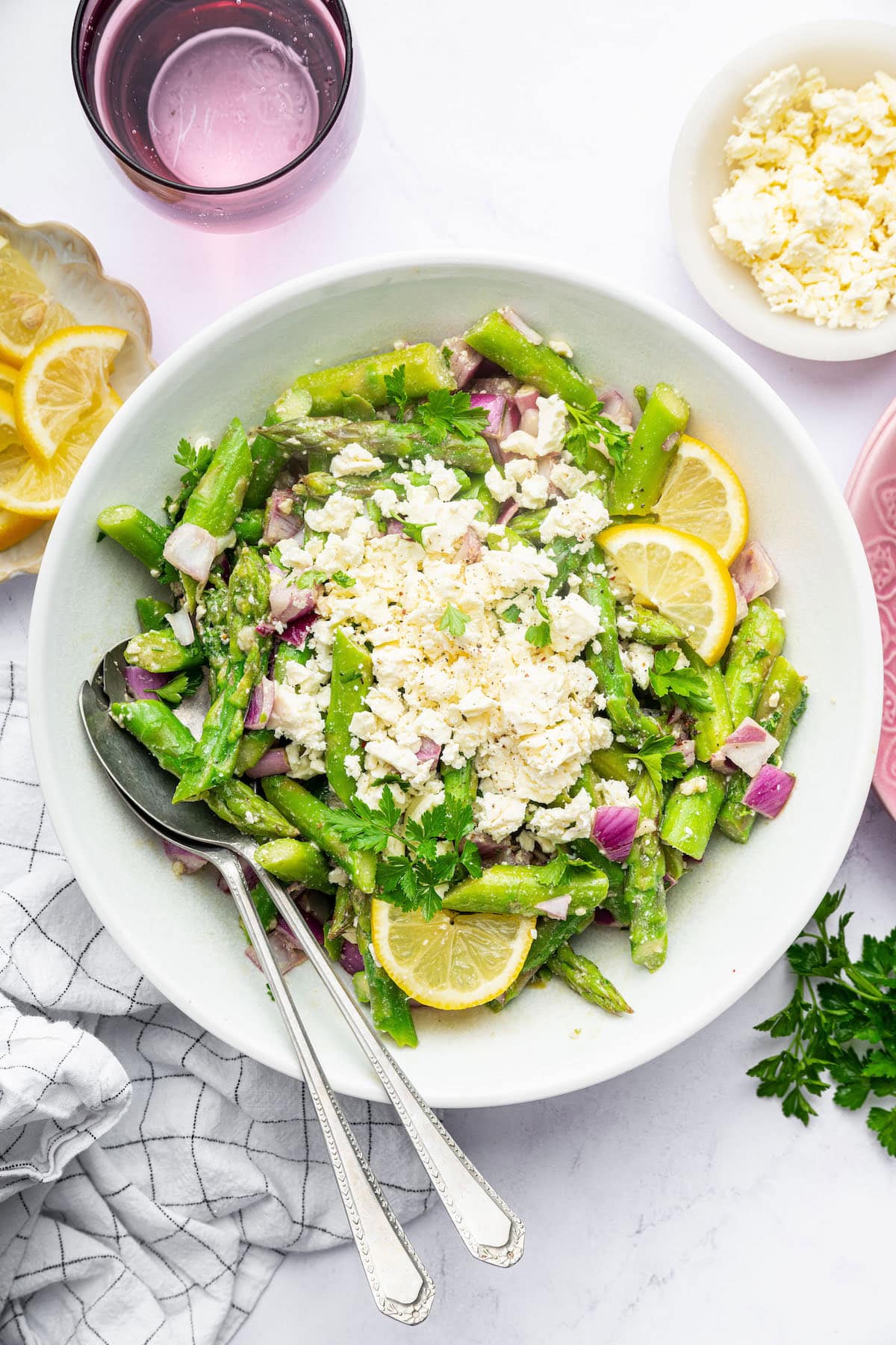 A large bowl of asparagus salad topped with crumbled feta and lemon slices, with serving utensils and fresh herbs on the side.