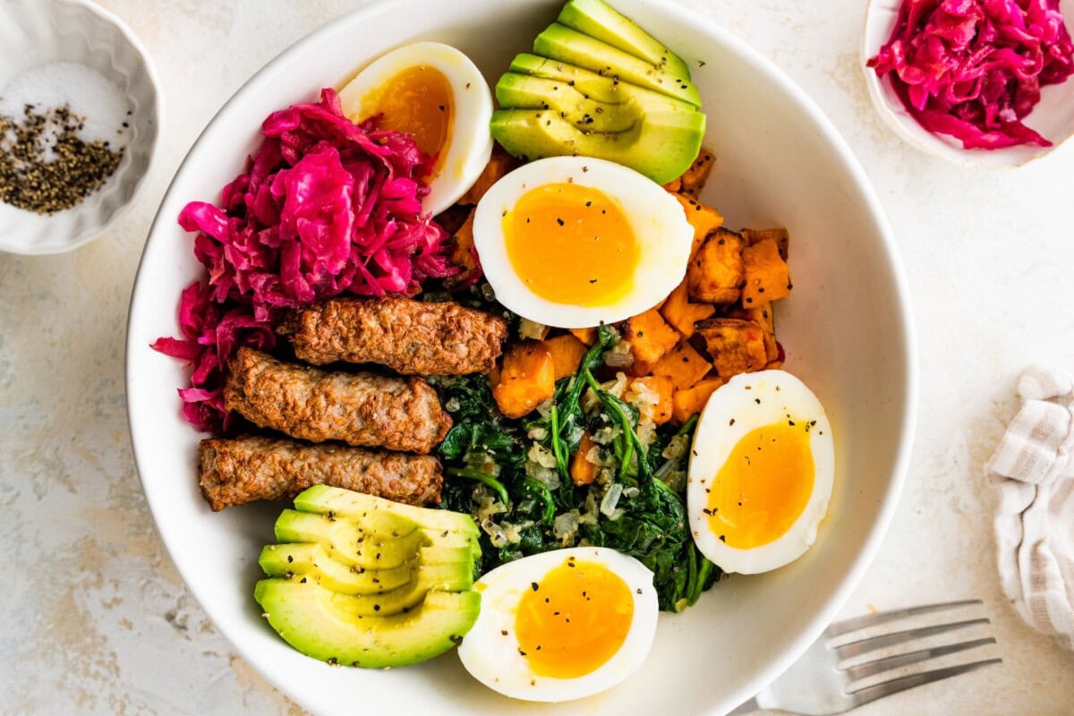 A bowl with two hard boiled eggs, avocado slices, turkey sausages, sweet potatoes, kale, and sauerkraut.