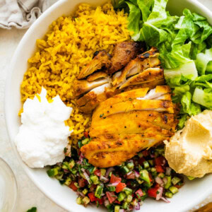 A large bowl serving cooked chicken, turmeric rice, lettuce, shirazi salad, hummus, and toum.