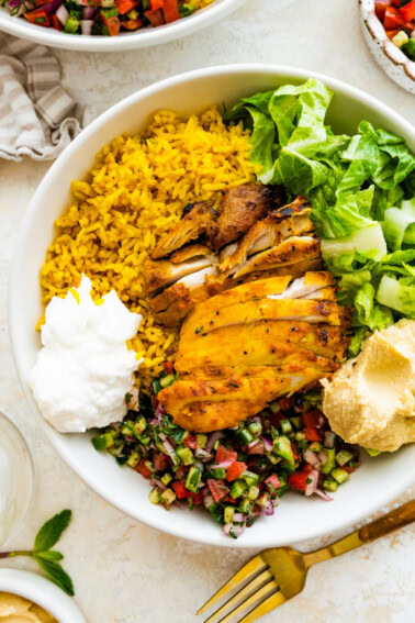 A large bowl serving cooked chicken, turmeric rice, lettuce, shirazi salad, hummus, and toum.