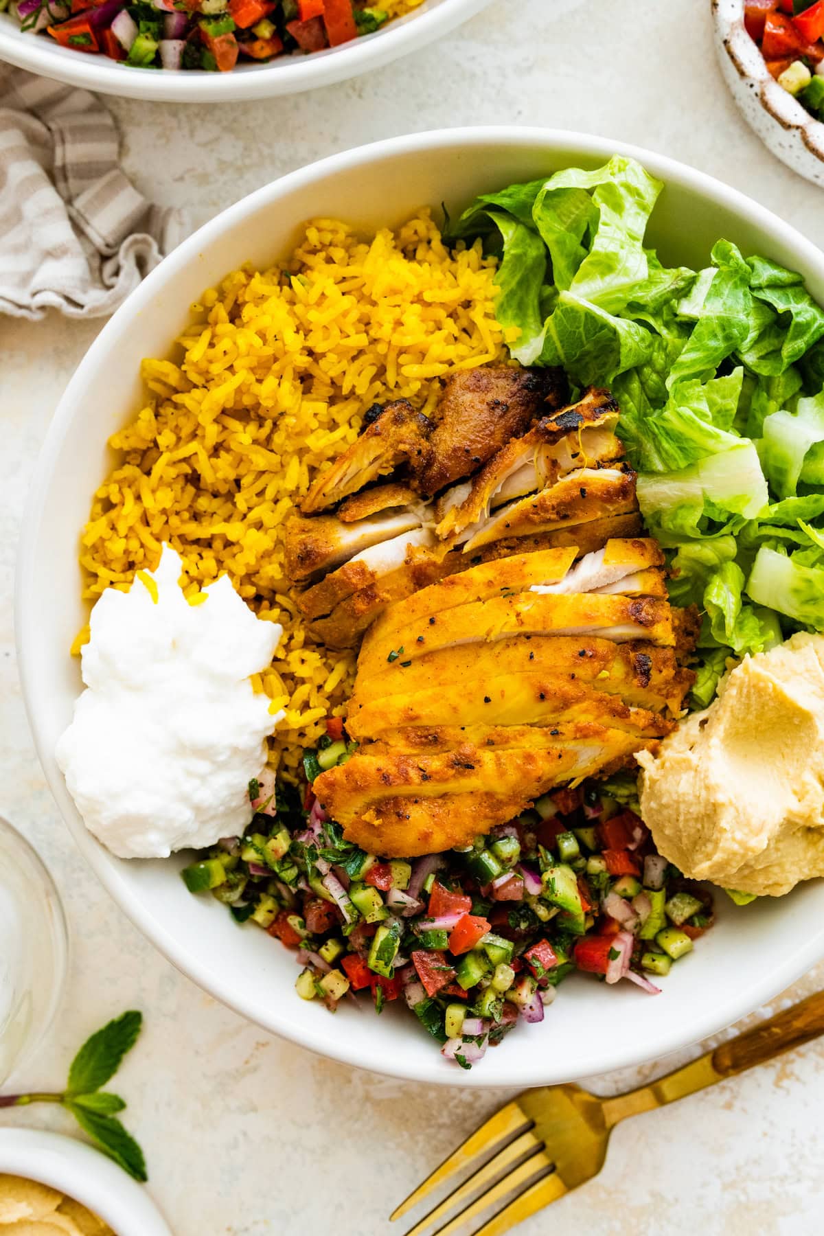 A large bowl serving cooked chicken, turmeric rice, lettuce, shirazi salad, hummus, and toum.