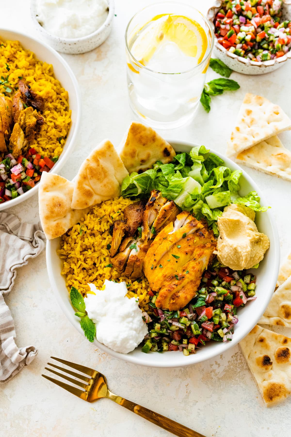 A chicken shawarma bowl with turmeric rice, romaine lettuce, hummus, shirazi salad, toum, and fresh pita bread on the side.