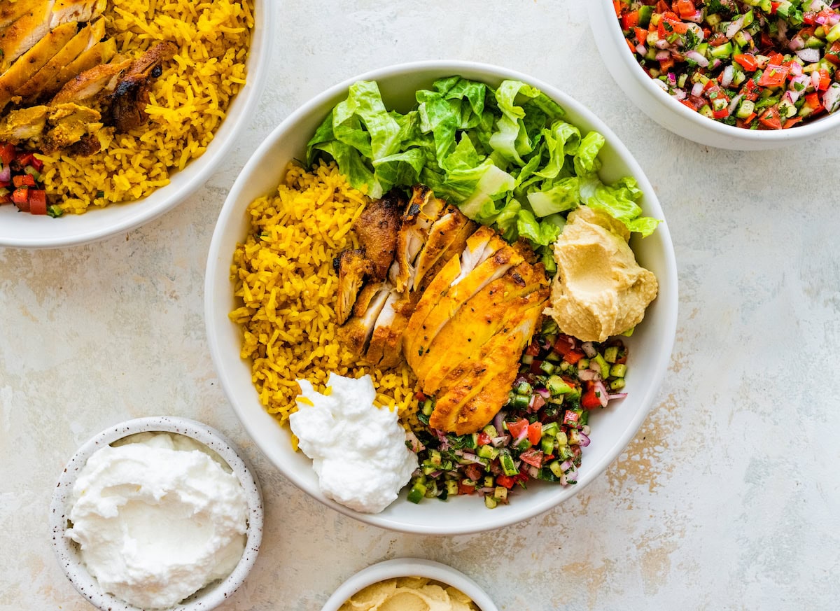 A large bowl serving chicken shawarma with rice, lettuce, shirazi salad, hummus, and toum.