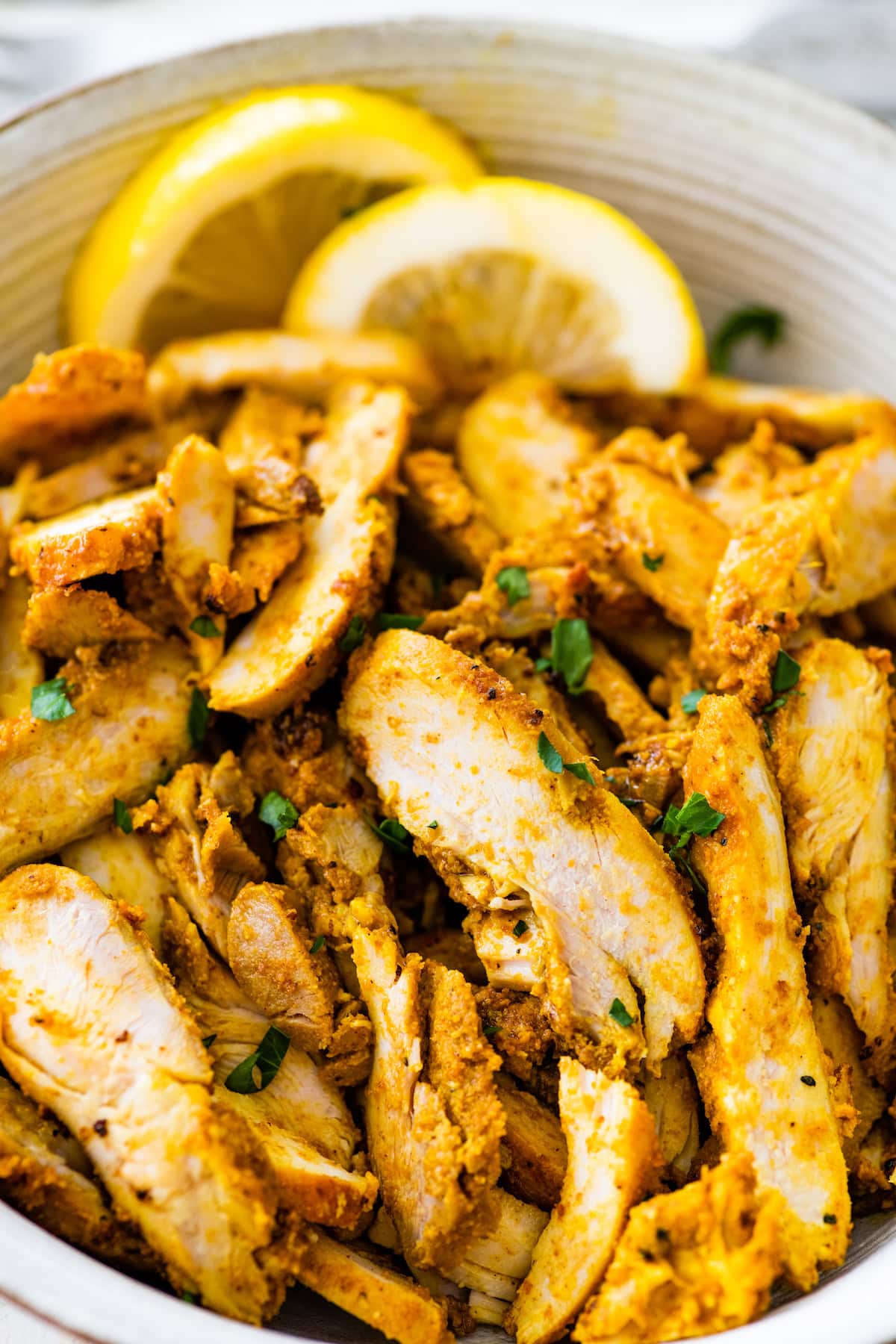 Sliced chicken shawarma in a bowl that's topped with fresh parsley. There are two lemon slices next to the chicken.