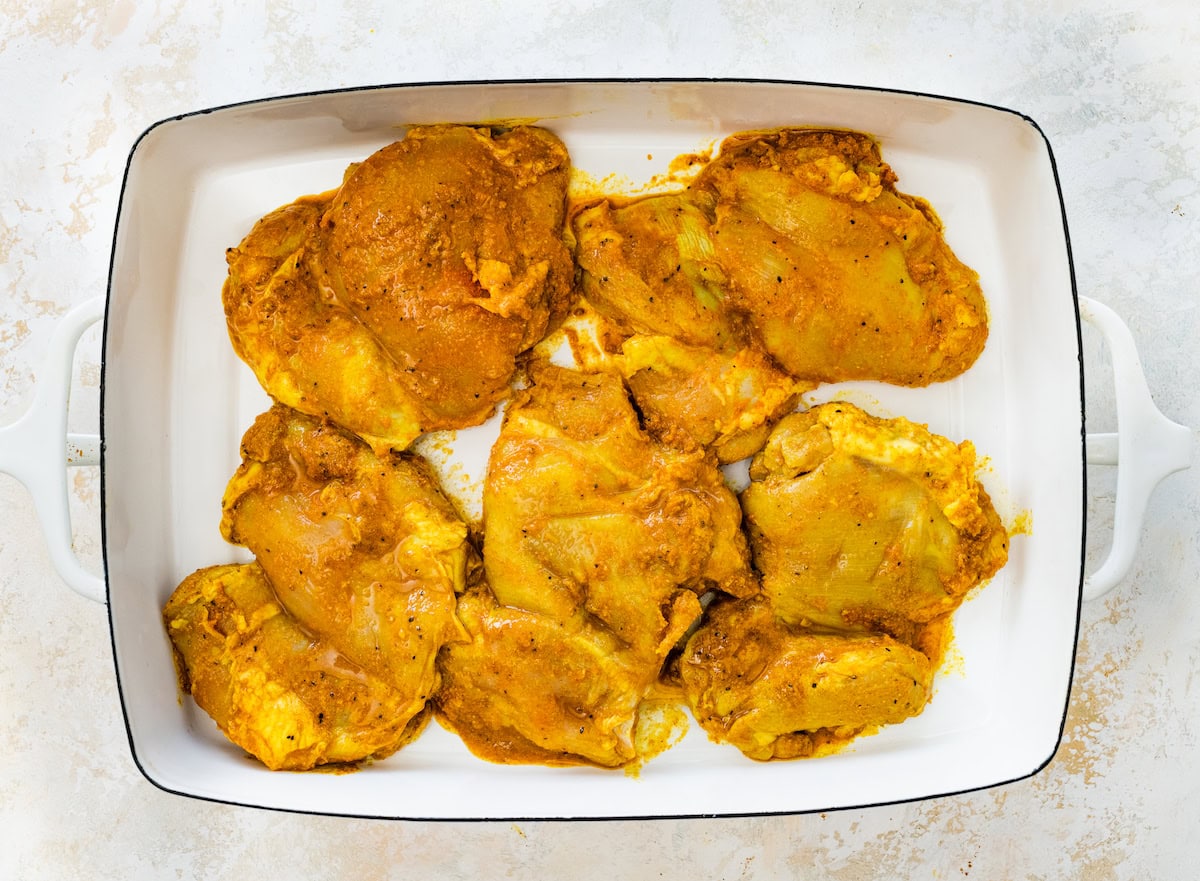 Five raw chicken thighs coated in the chicken shawarma marinade in a white baking dish.