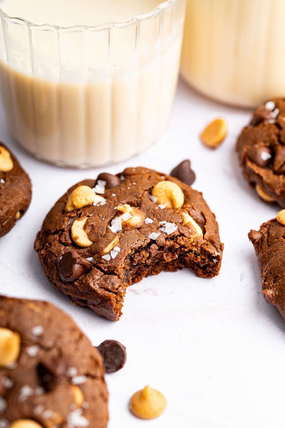 One chocolate peanut butter cookie with a bite taken out of it. The cookie is topped with flaky sea salt and there is a glass of milk in the background.