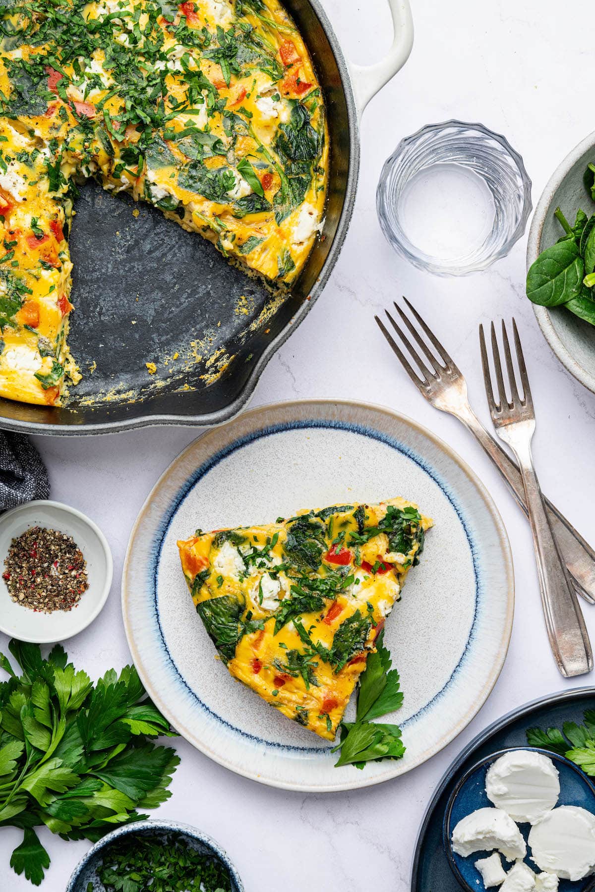 A frittata slice served on a plate next to two silver forks. The full frittata is in a cast iron skillet next to the plate.