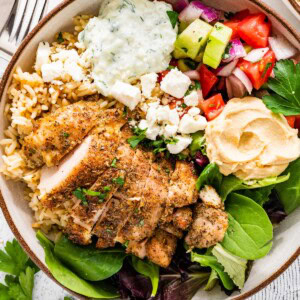 A bowl served with cooked chicken, mixed greens, tzatziki, hummus, feta cheese, and a cucumber tomato salad.