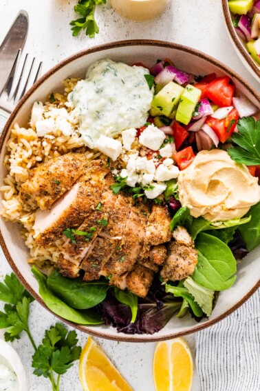 A bowl served with cooked chicken, mixed greens, tzatziki, hummus, feta cheese, and a cucumber tomato salad.