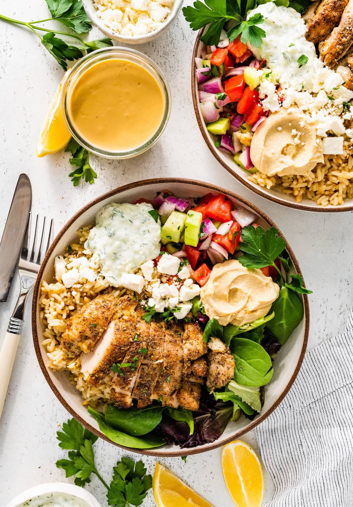 Two bowls served with mixed greens, Greek chicken, tzatziki, hummus, cucumber tomato salad and feta cheese. There is a tahini dressing next to the bowls.